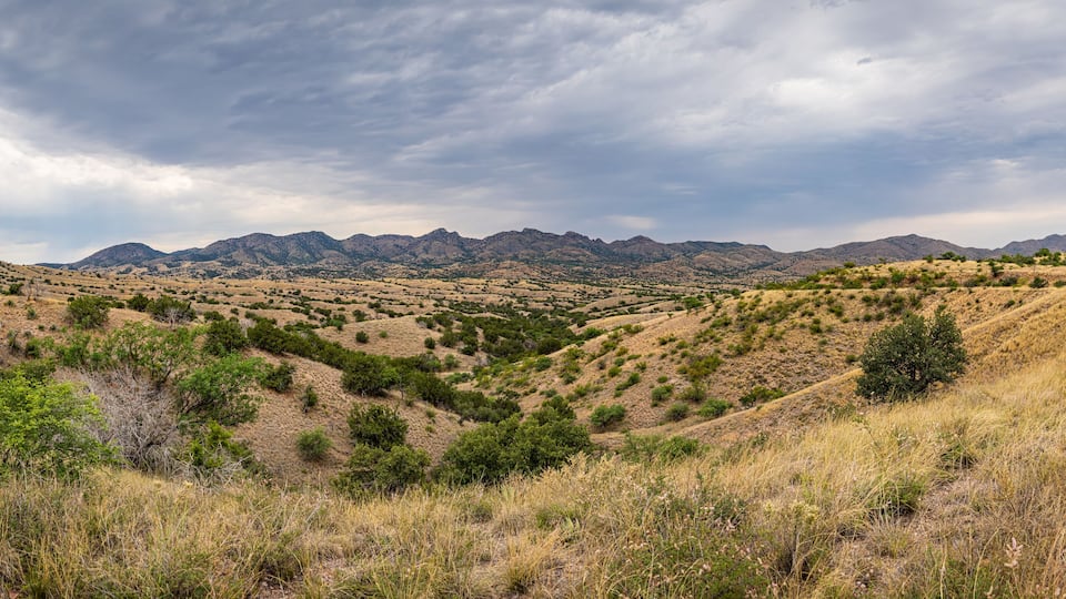 A dry, hilly landscape with sparse vegetation and distant mountains under a cloudy sky.