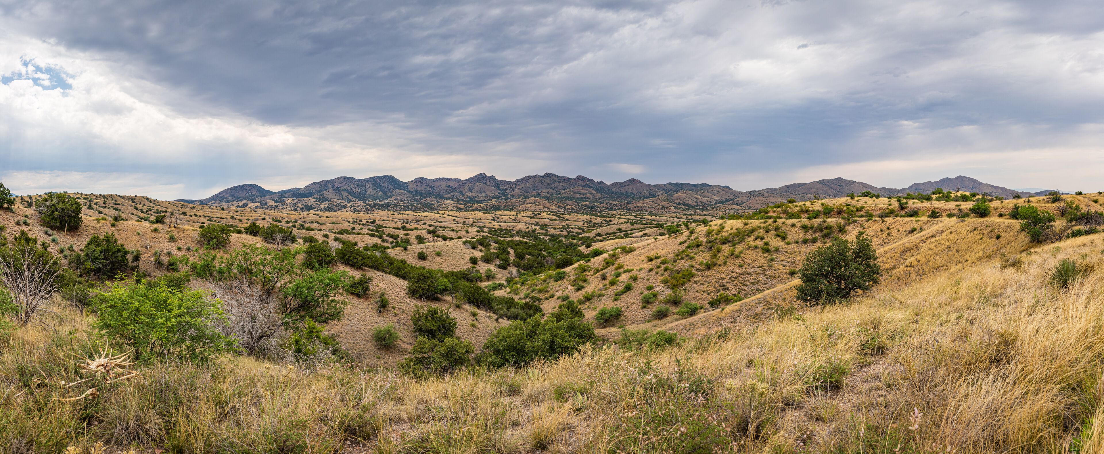 A dry, hilly landscape with sparse vegetation and distant mountains under a cloudy sky.
