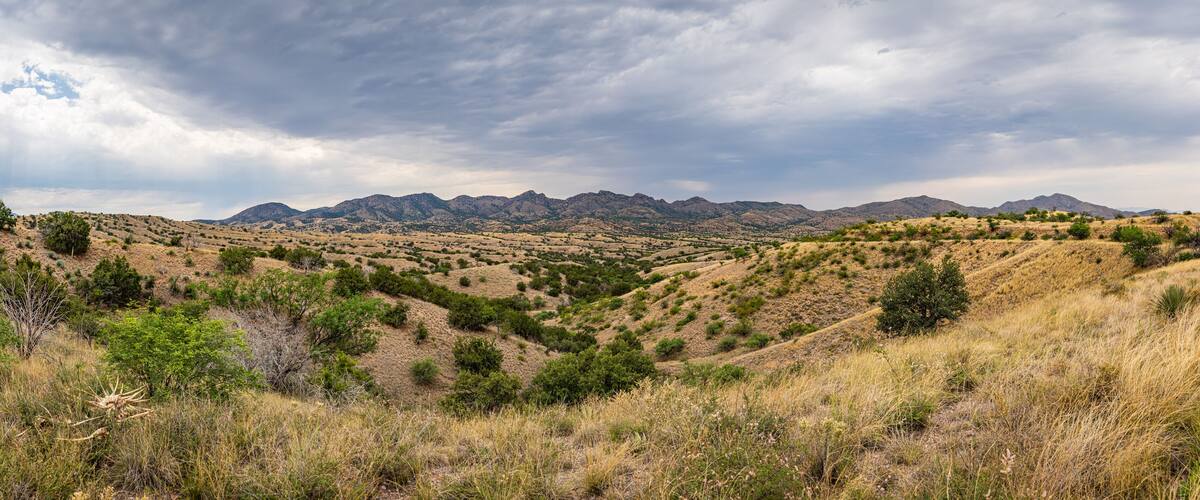 A dry, hilly landscape with sparse vegetation and distant mountains under a cloudy sky.