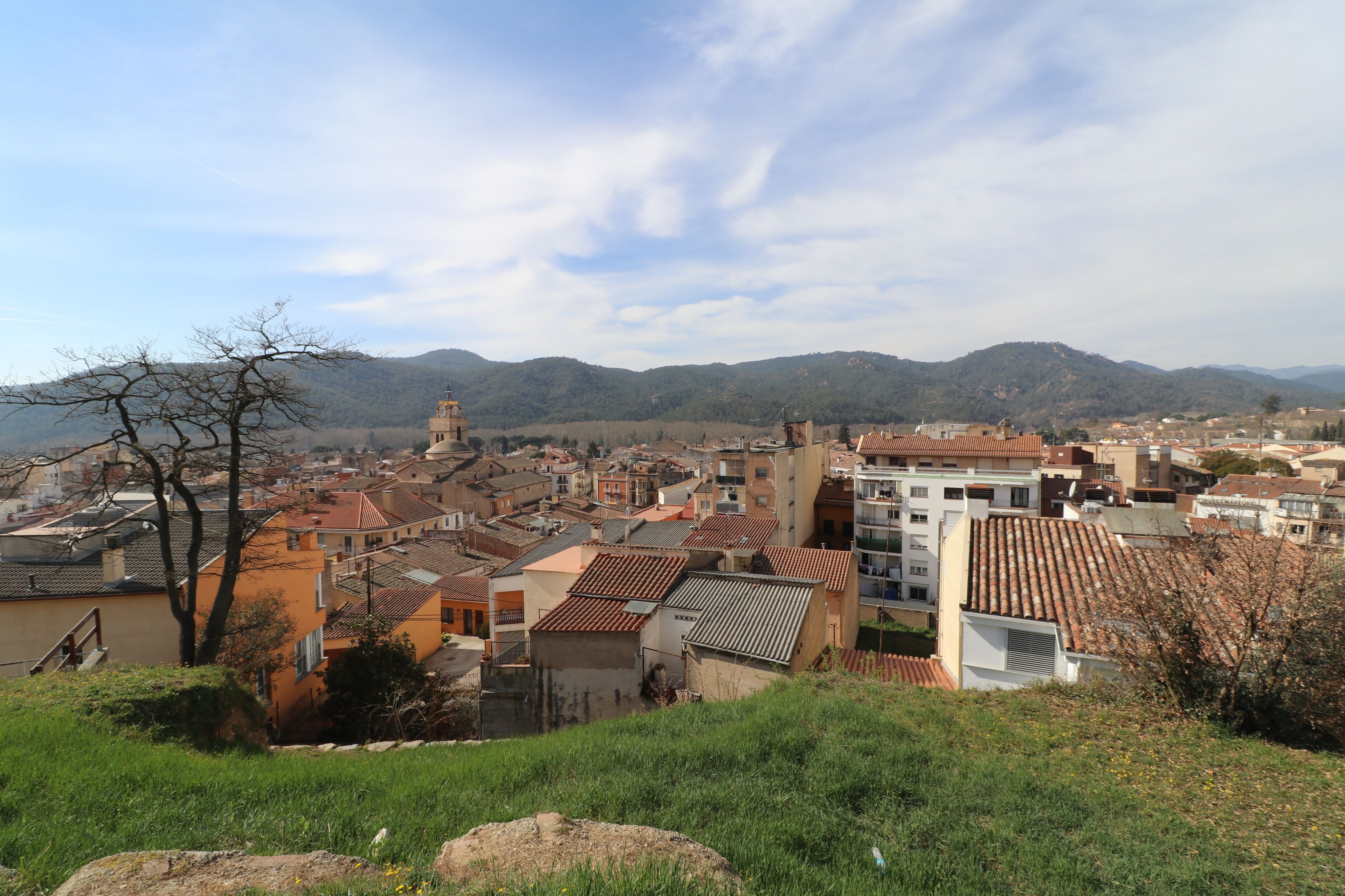 A town with terracotta-roofed buildings, a prominent church tower, and rolling hills in the background.