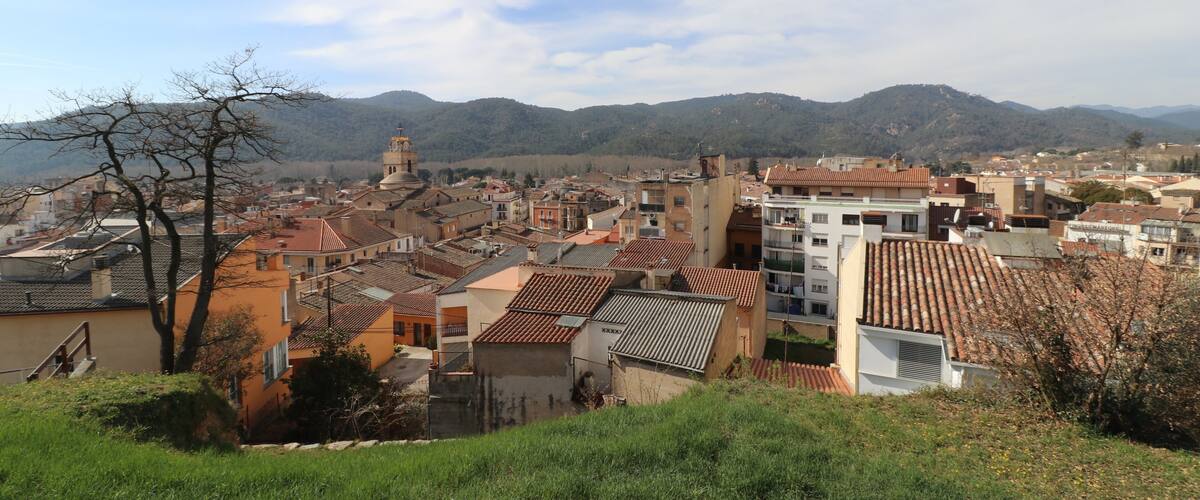 A town with terracotta-roofed buildings, a prominent church tower, and rolling hills in the background.
