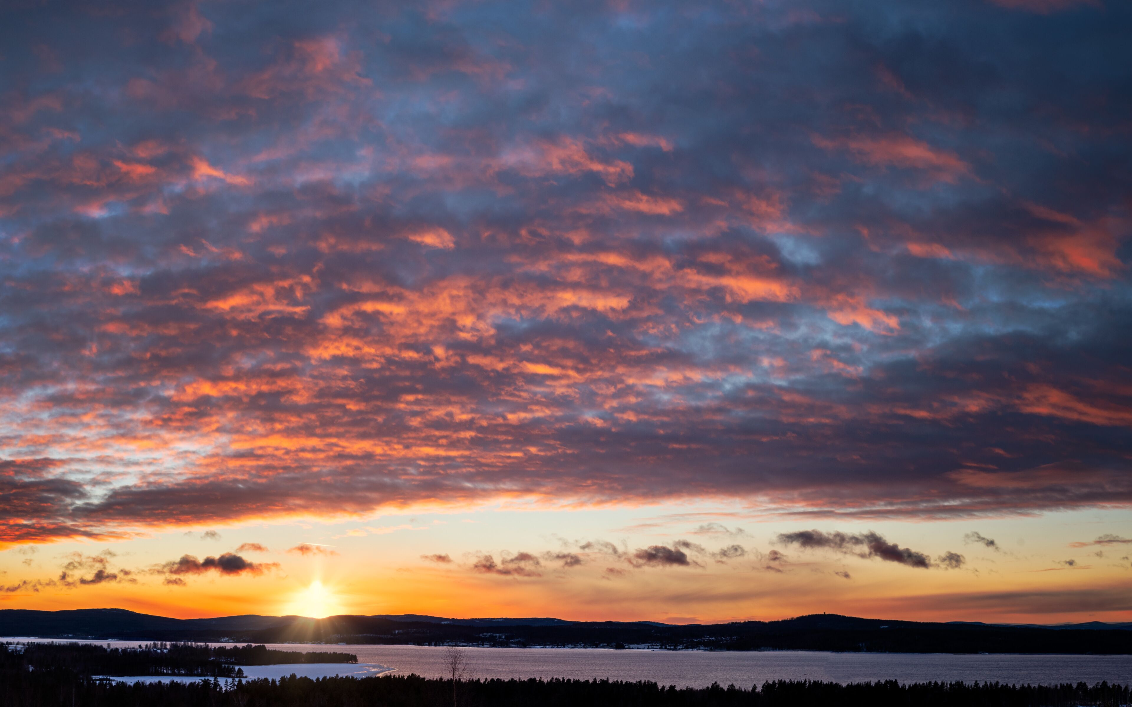 A sunset over a body of water with a forested shoreline and distant hills.