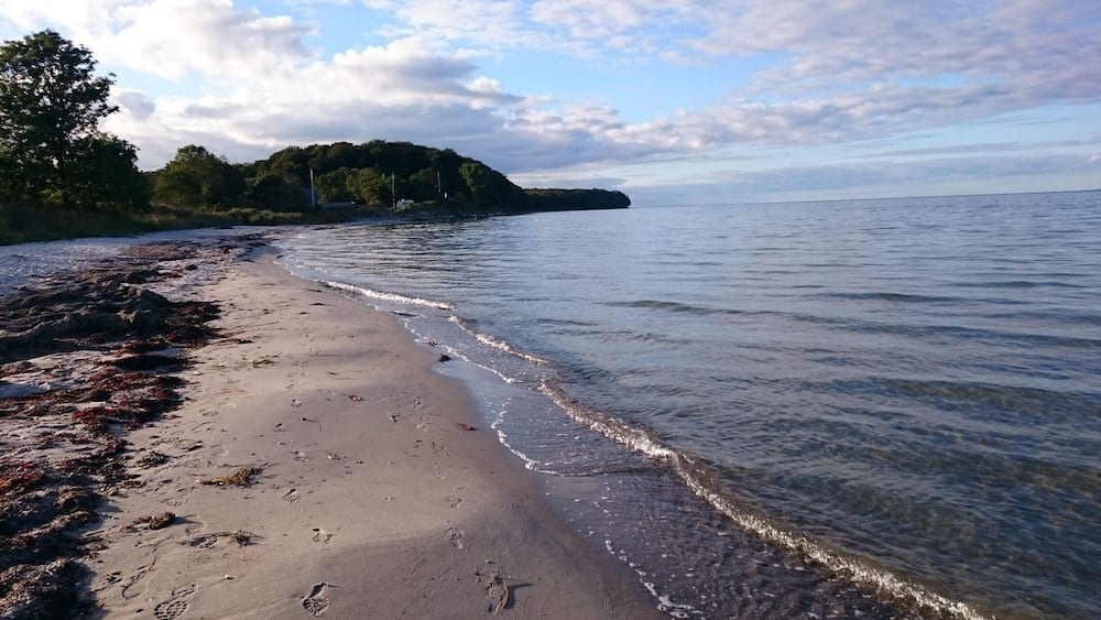 Ein sandiger Strand mit sanften Wellen, eine bewaldete Insel in der Ferne und ein teilweise bewölkter Himmel.