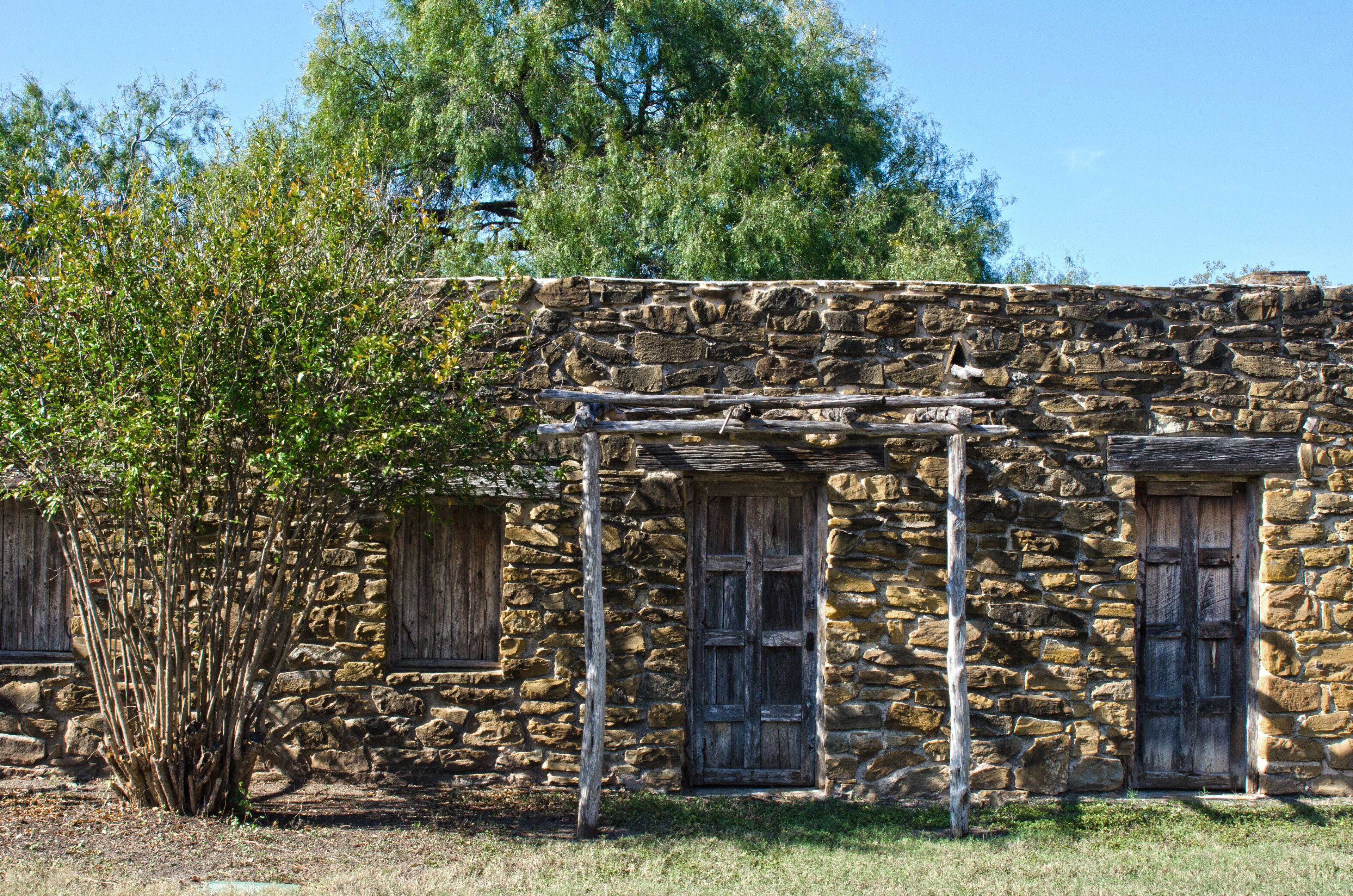 A stone building with wooden doors and windows, surrounded by trees and grass.