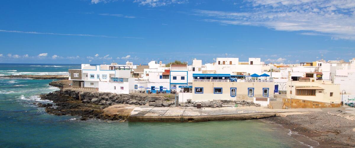 A coastal town with white buildings and blue accents, a rocky shoreline, and the ocean in the background.