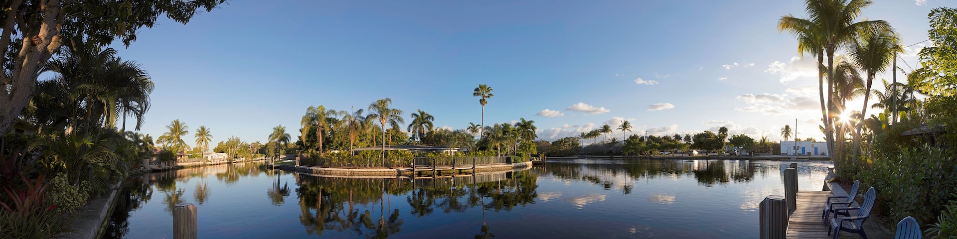 A calm lake with a wooden dock and chairs, surrounded by palm trees and a clear sky.
