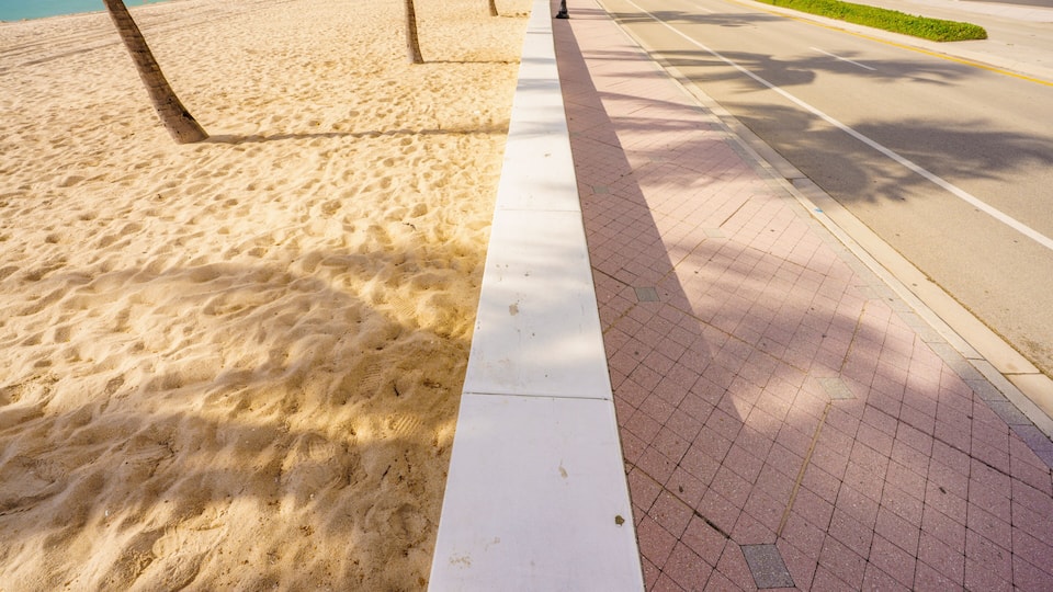 A beach with sand, a white line, and a paved sidewalk.