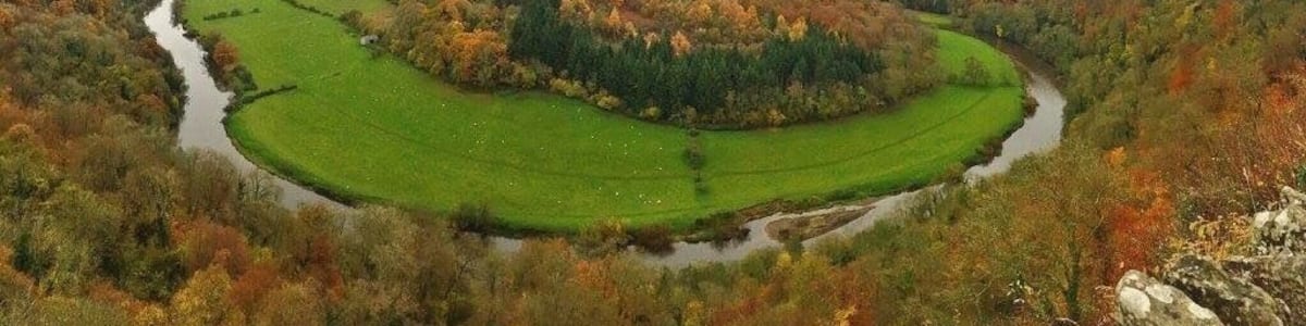 Ein Fluss schlängelt sich durch ein Tal mit herbstlich gefärbten Bäumen und grünen Feldern.