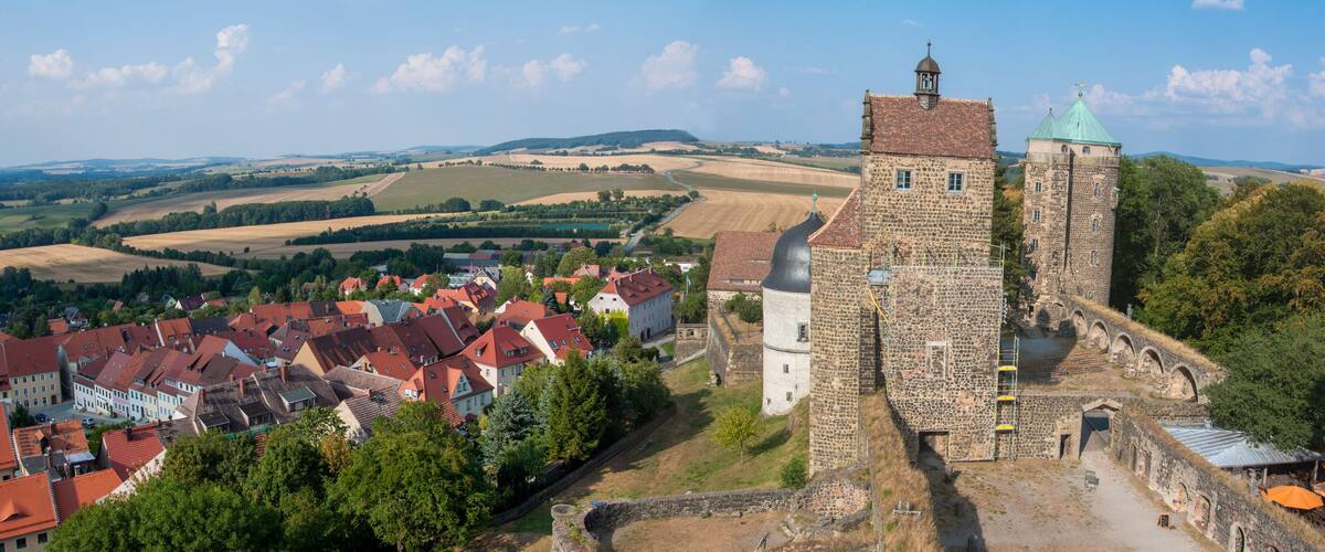 A historic tower with a green roof, surrounded by a village with red-roofed houses and fields in the background.