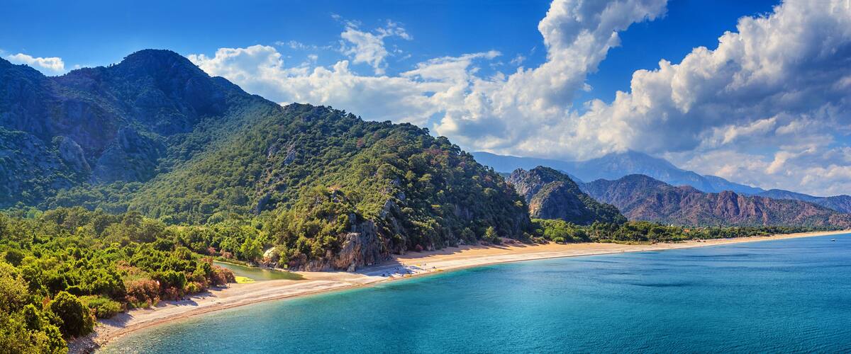 A coastal landscape with a beach, clear blue waters, and a mountainous backdrop.