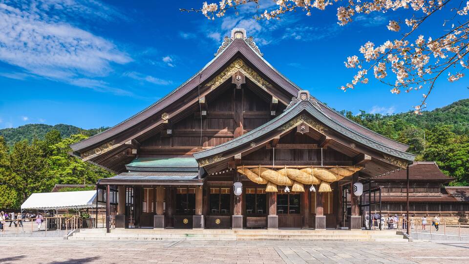 A traditional wooden building with a steeply pitched roof, surrounded by a paved area and cherry blossoms.