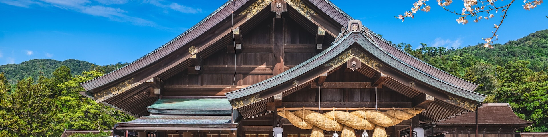 A traditional wooden building with a steeply pitched roof, surrounded by a paved area and cherry blossoms.