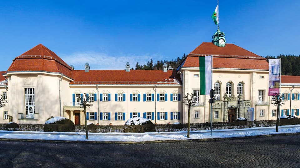 A large, historic building with a red roof and multiple windows, surrounded by snow and trees.