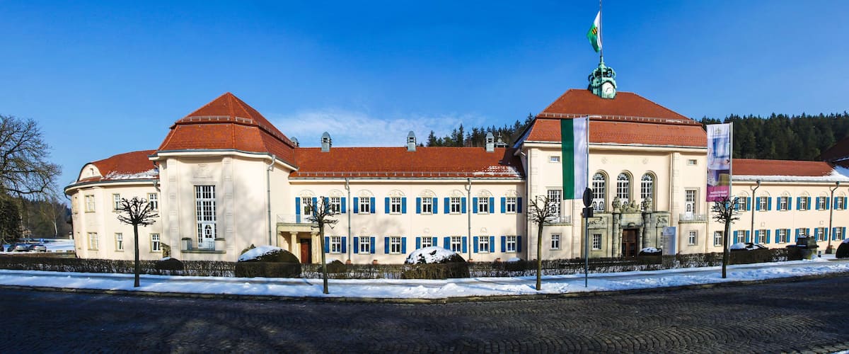 A large, historic building with a red roof and multiple windows, surrounded by snow and trees.