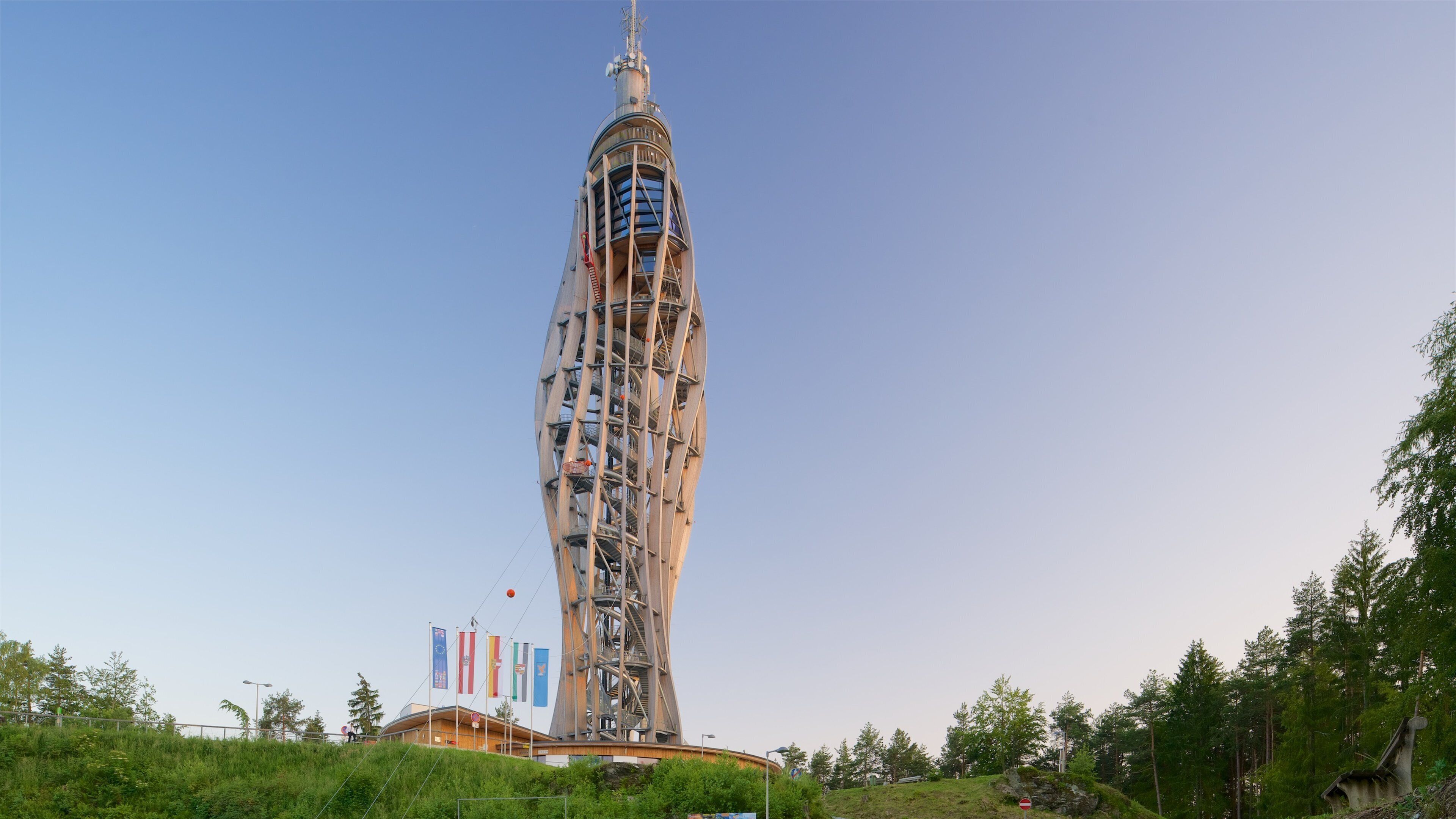 A tall, spiraling tower with a unique architectural design, surrounded by greenery and flags.