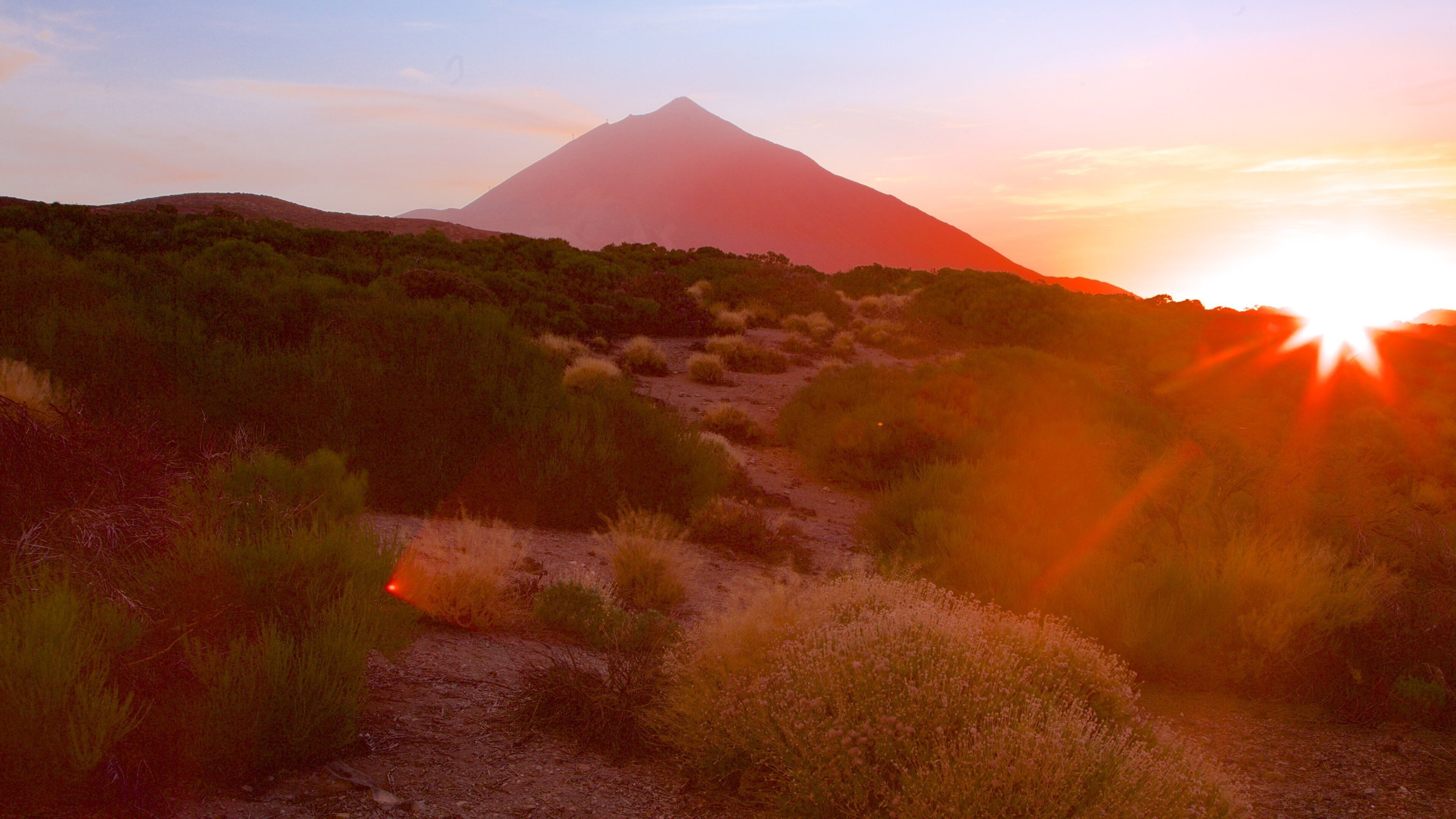 A desert landscape with a mountain in the background and the sun setting.