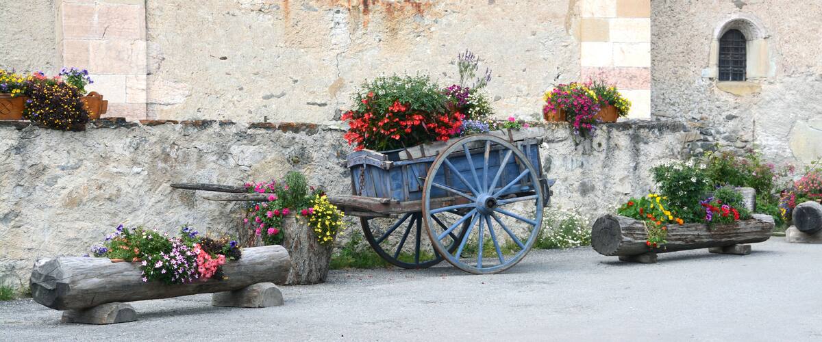 A rustic stone wall with a blue cart filled with colorful flowers, surrounded by wooden logs and planters.