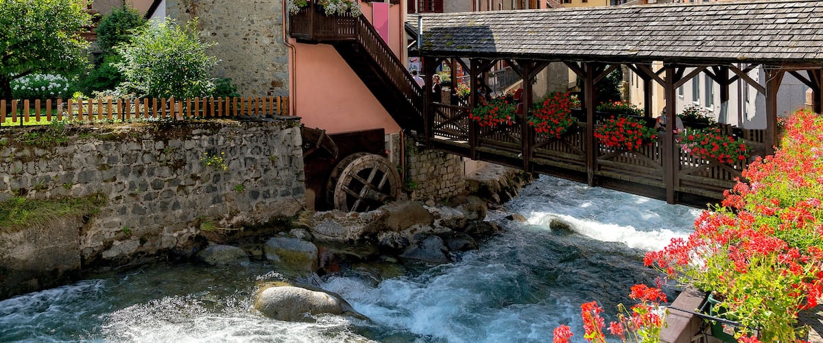 A stone bridge over a flowing river, surrounded by colorful buildings and a mountainous backdrop.