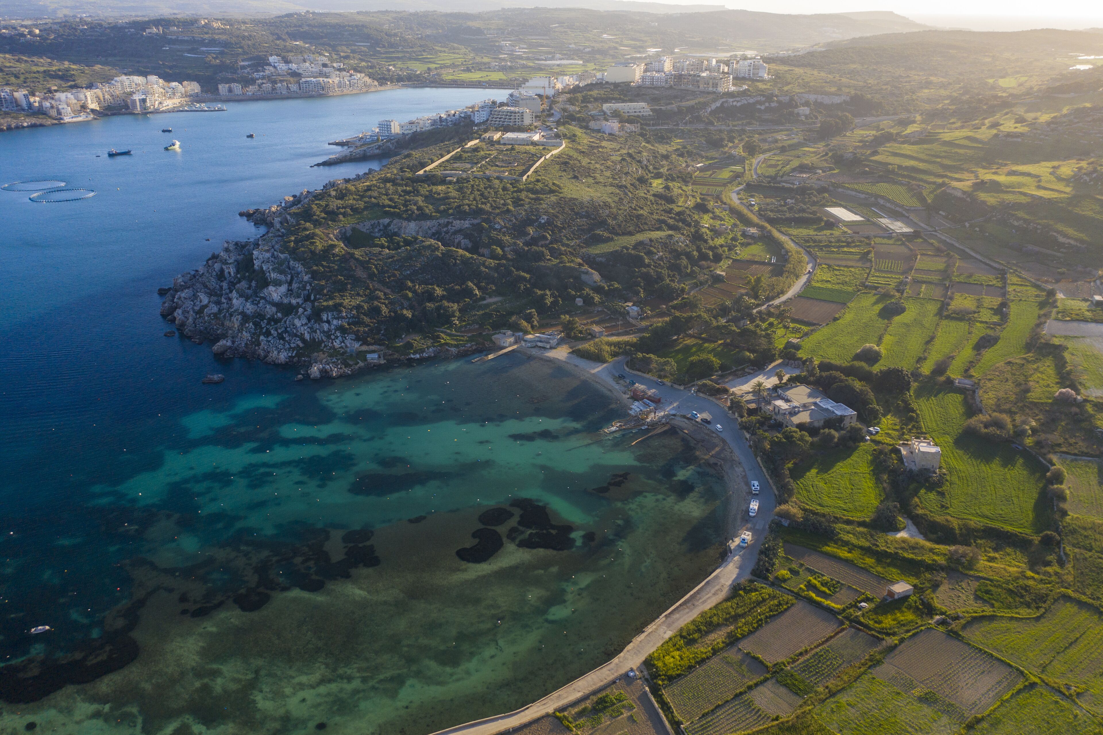 An aerial view of a coastal area with a clear road, green fields, and a body of water with boats.