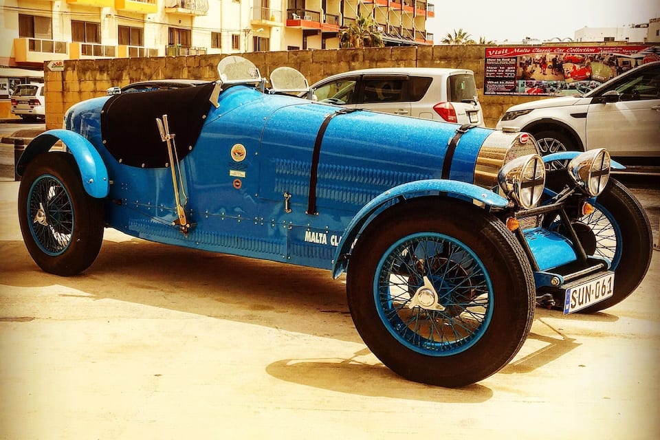 Un coche deportivo azul antiguo estacionado en una calle con edificios al fondo.