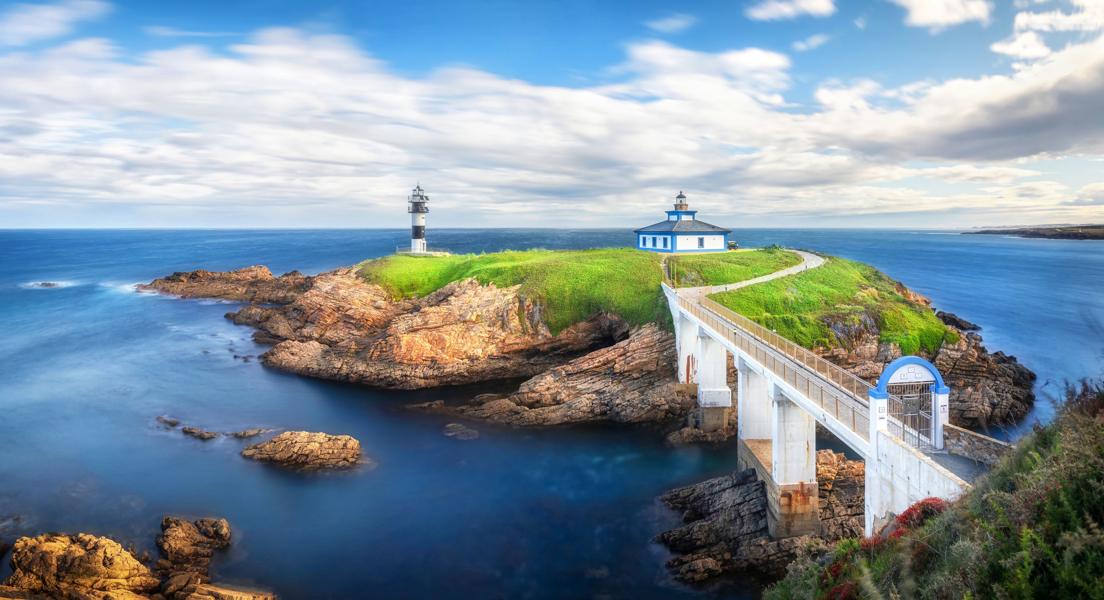 A coastal landscape with a lighthouse, a bridge, and a building on a grassy hill.