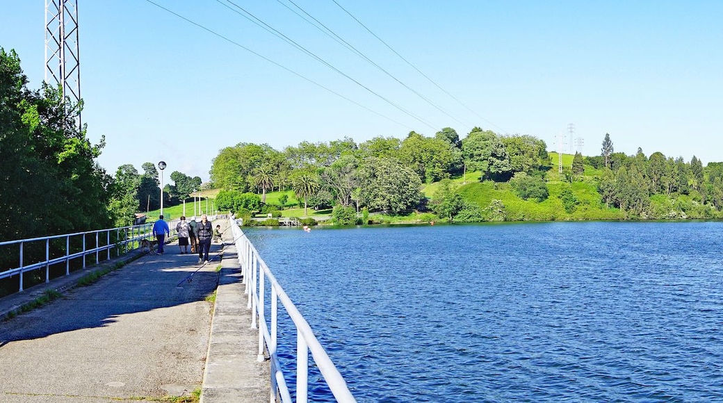 Ein großes Gewässer mit einer Brücke auf der linken Seite, umgeben von Grünflächen und Hügeln im Hintergrund.