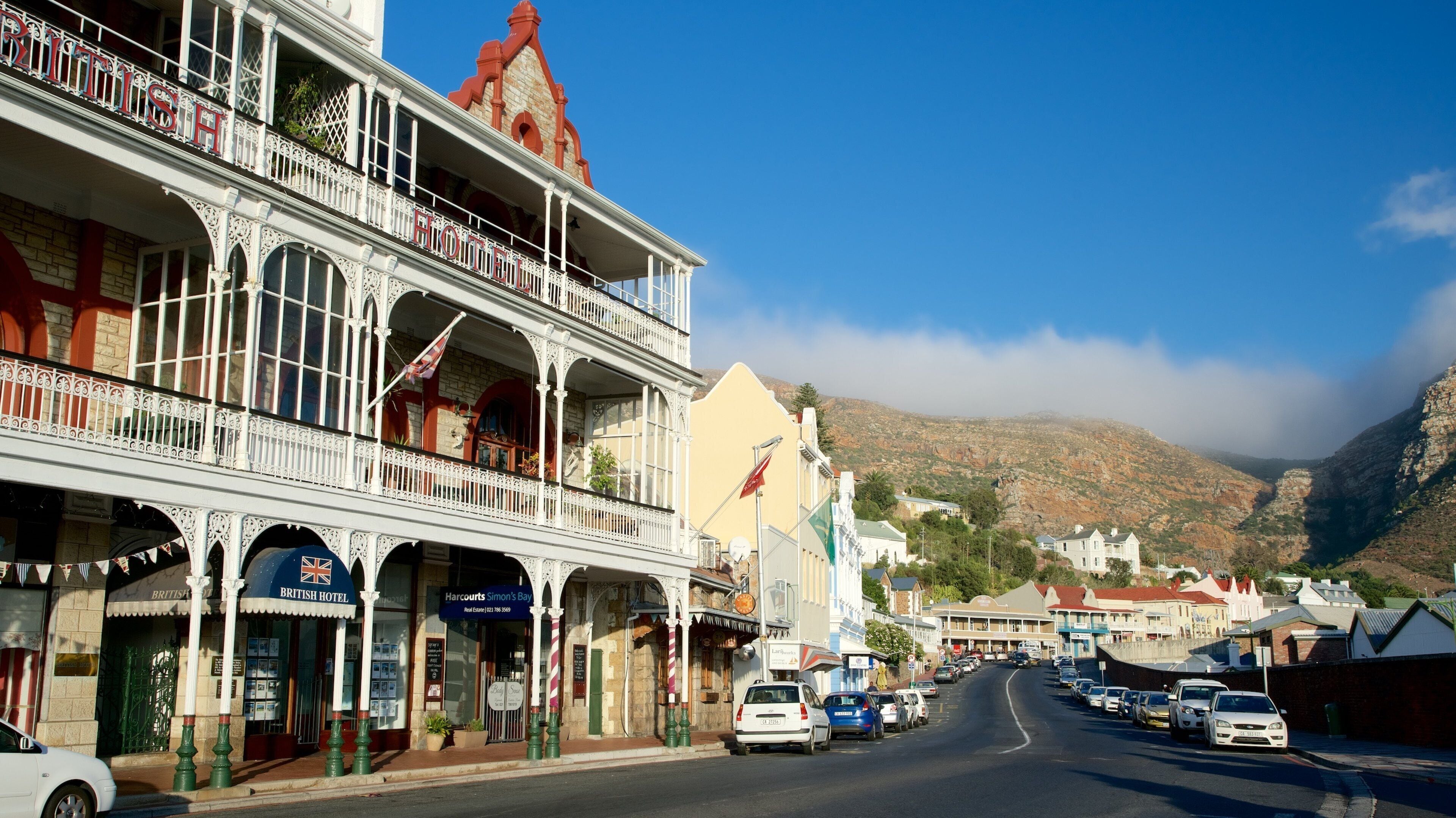 A street with British Hotel and Harbourside Hotel, cars parked along the road, and mountains in the background.