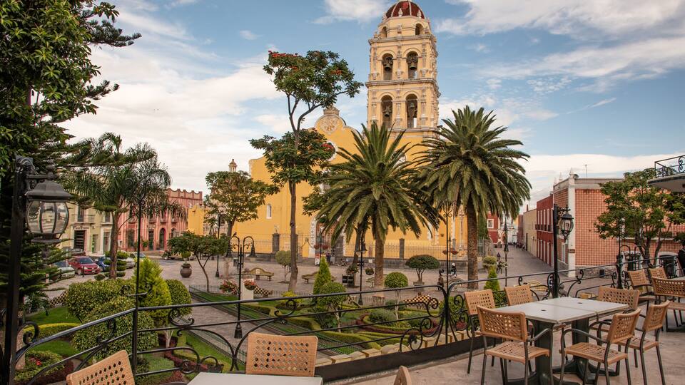 A plaza with a yellow building, palm trees, and a bell tower.