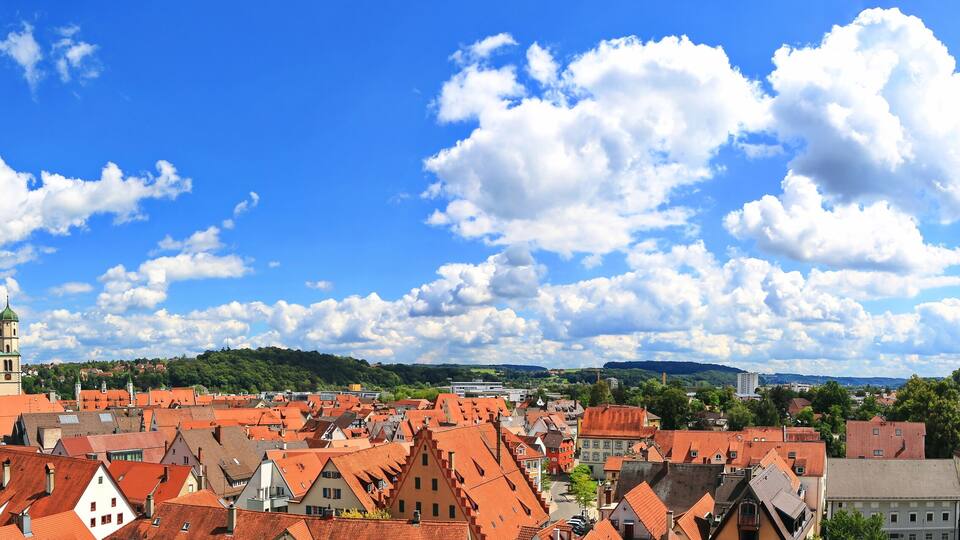 A town with red-roofed buildings, a prominent church tower, and a clear blue sky with scattered clouds.