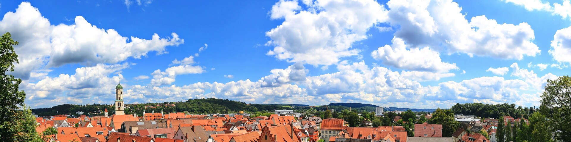 A town with red-roofed buildings, a prominent church tower, and a clear blue sky with scattered clouds.