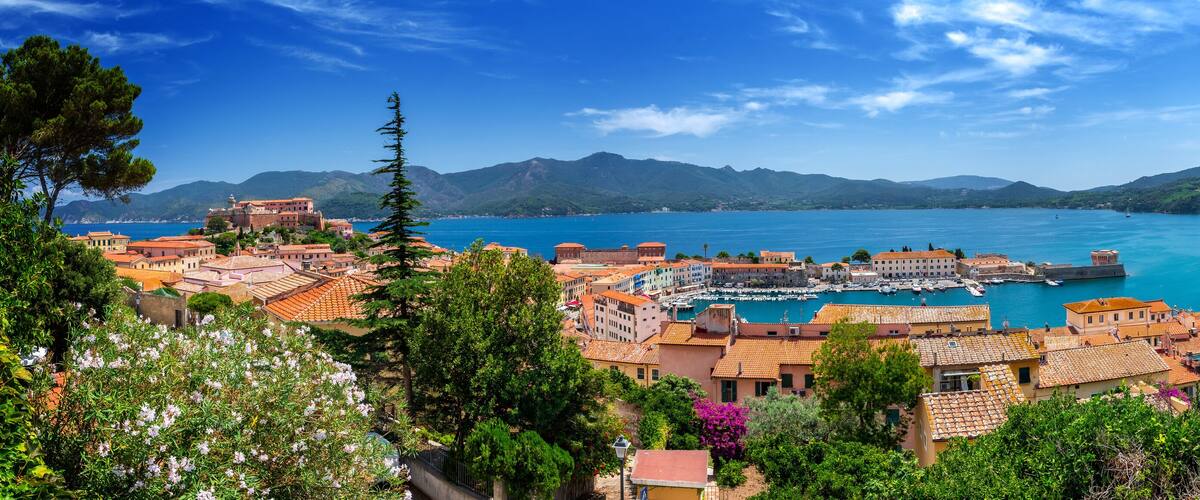 A coastal town with terracotta-roofed buildings, a marina, and mountains in the background.