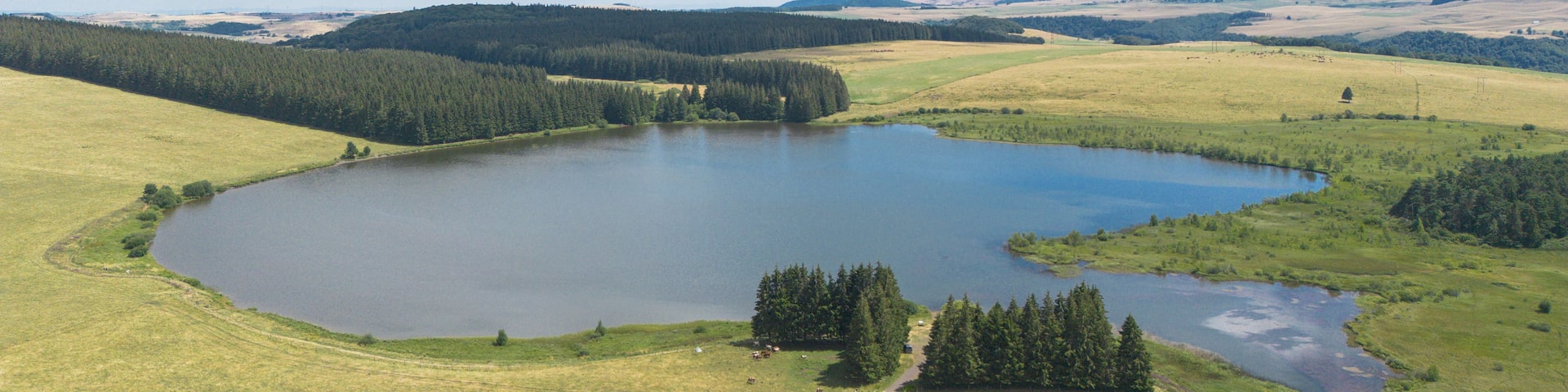 A lake surrounded by forested hills and open fields under a partly cloudy sky.