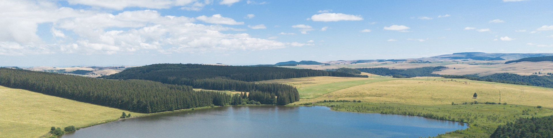 Un lago rodeado de colinas boscosas y campos abiertos bajo un cielo parcialmente nublado.