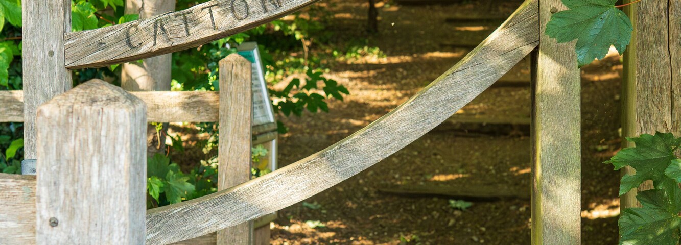 Rustic wooden gate entrance to Carlton Park with weathered fence leading to forest trail. Charming country park gateway with natural woodland path, green foliage and dappled sunlight background.
