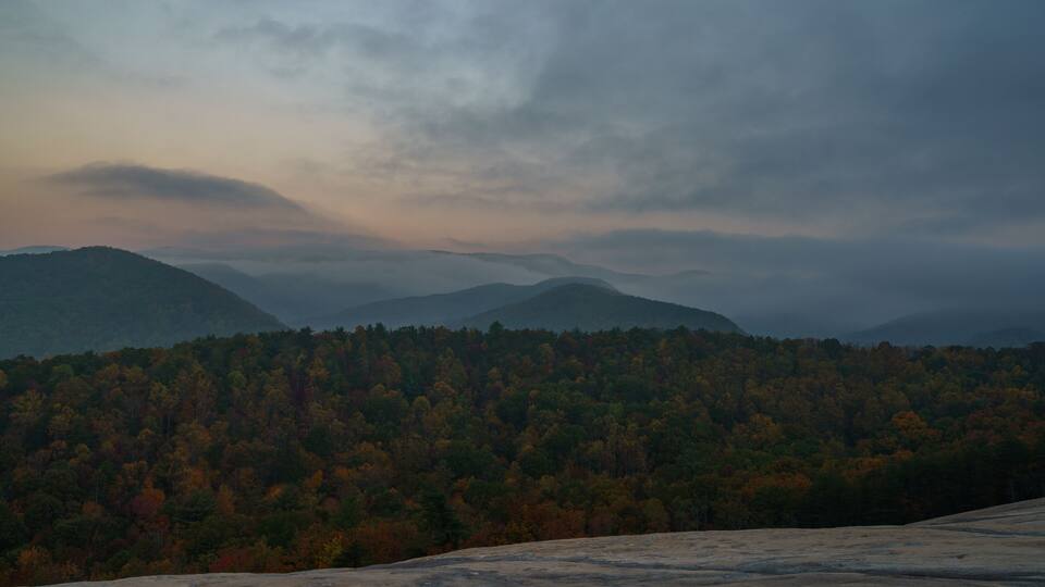 A mountainous landscape with autumn-colored trees and a cloudy sky.