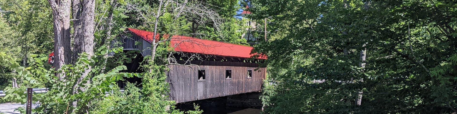 A wooden covered bridge with a red roof, surrounded by lush greenery and trees.