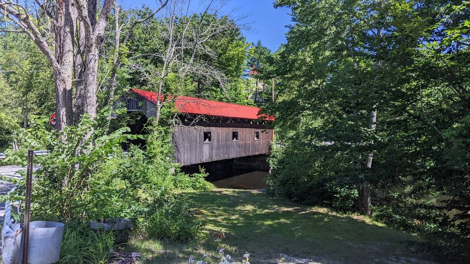 A wooden covered bridge with a red roof, surrounded by lush greenery and trees.