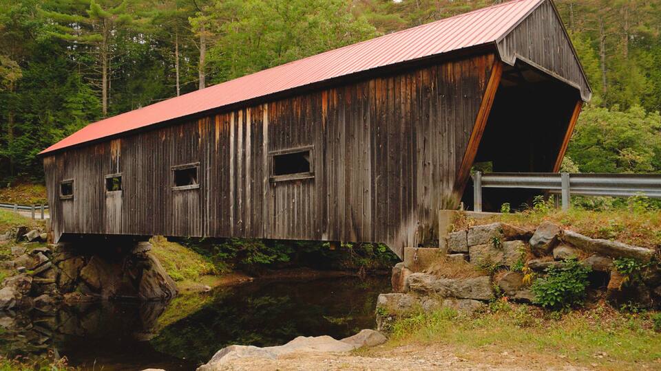 A wooden covered bridge over a stream, surrounded by trees and rocks.