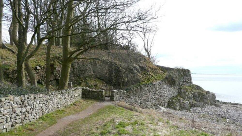 Ein Küstenpfad mit einer Steinmauer, Bäumen und Blick aufs Meer.