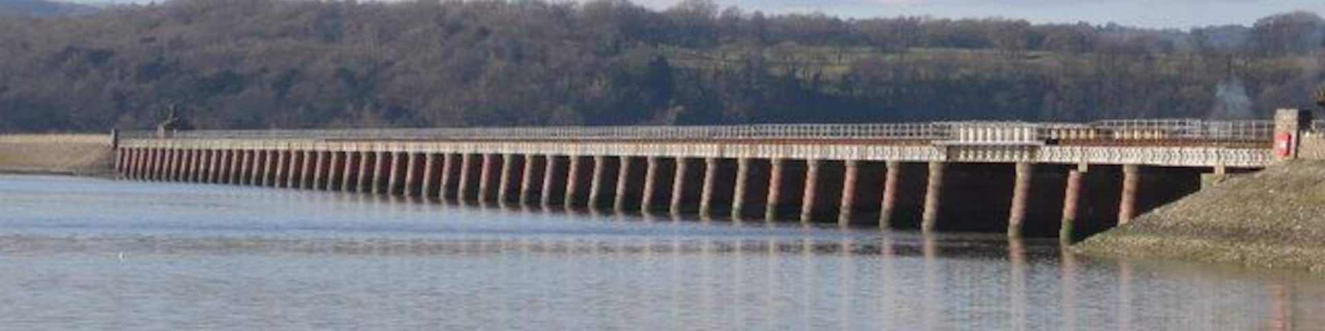 A long bridge over a calm river with grassy banks and a forested hill in the background.