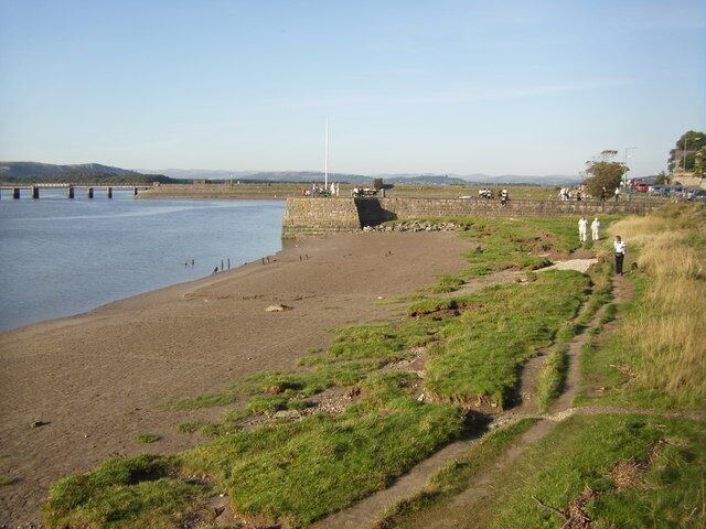A riverside path with a bridge in the background and a grassy area with people walking.