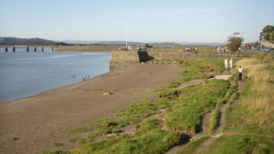A riverside path with a bridge in the background and a grassy area with people walking.