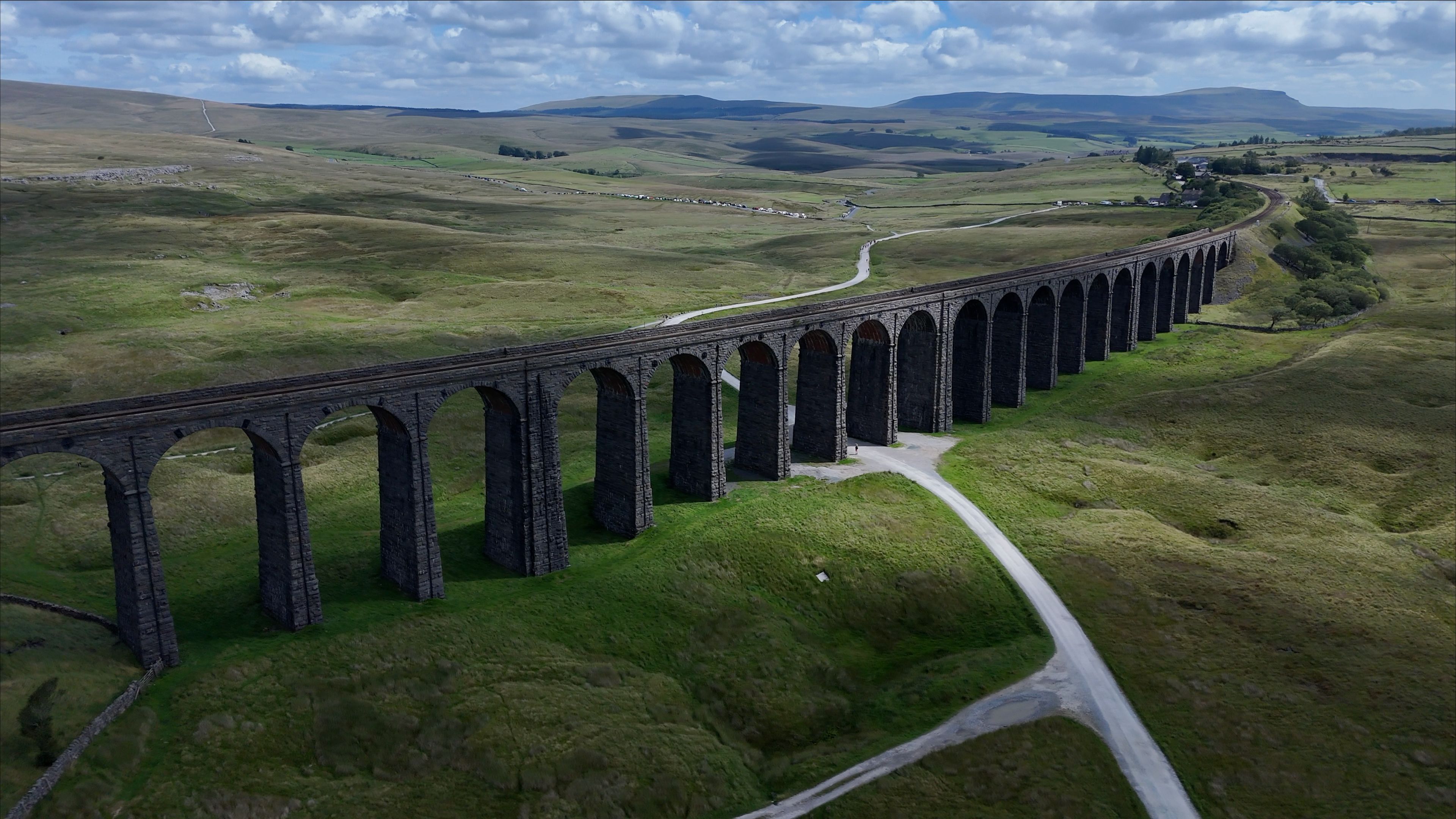 Aerial view of Ribblehead Viaduct, a historic stone railway bridge spanning a green valley with rolling hills, a winding road, and clouds in Carnforth, England, United Kingdom.