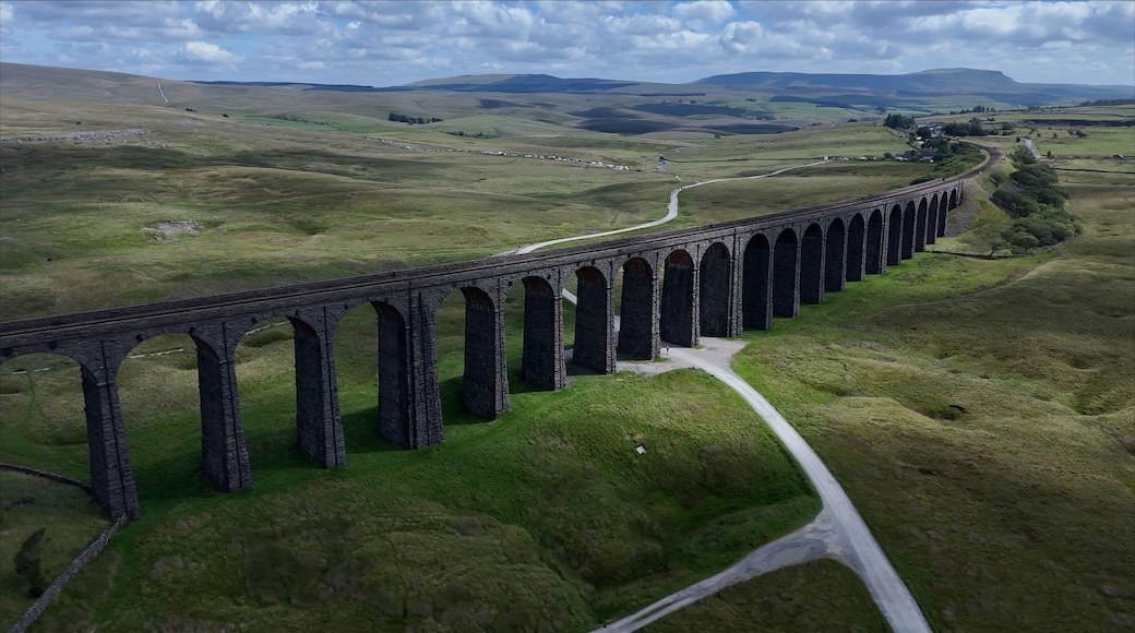 Aerial view of Ribblehead Viaduct, a historic stone railway bridge spanning a green valley with rolling hills, a winding road, and clouds in Carnforth, England, United Kingdom.