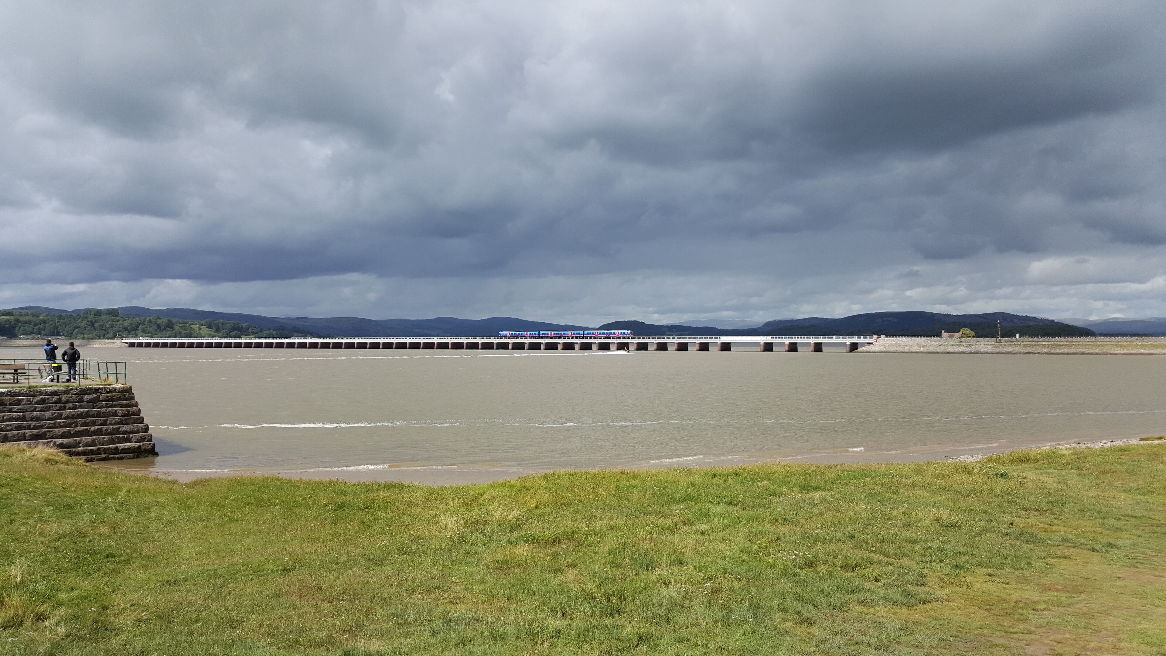 A river with a bridge, grassy banks, and distant mountains under a cloudy sky.