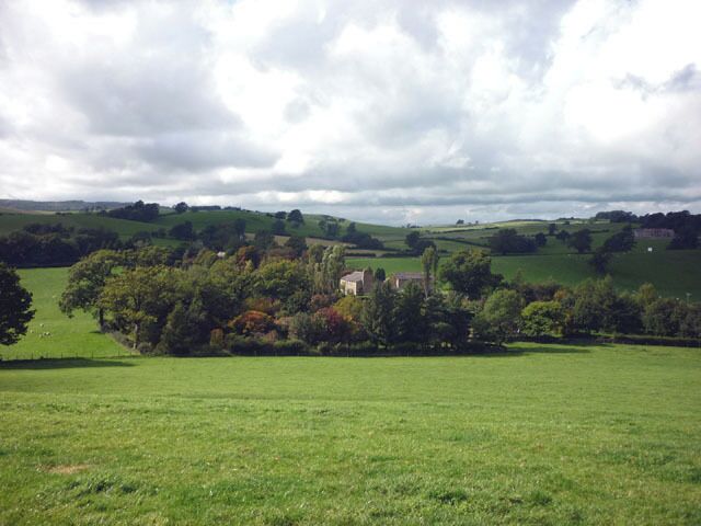 A rural landscape with rolling hills, a house surrounded by trees, and a grassy field.