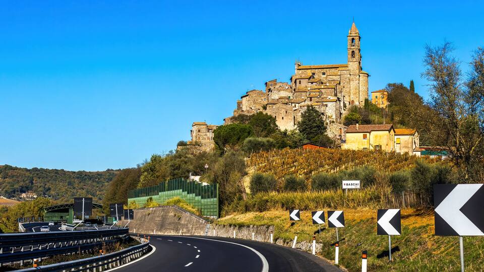 A winding road with directional signs, a historic stone building on a hill, and a clear blue sky.