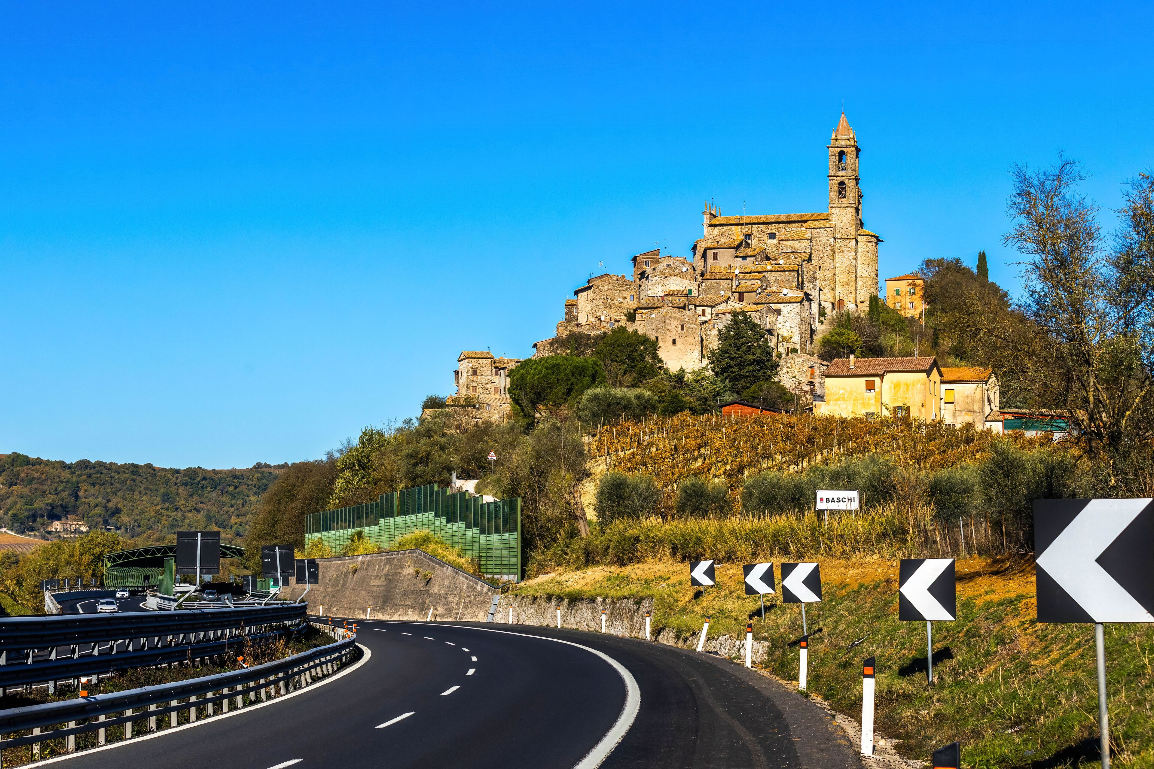 A winding road with directional signs, a historic stone building on a hill, and a clear blue sky.