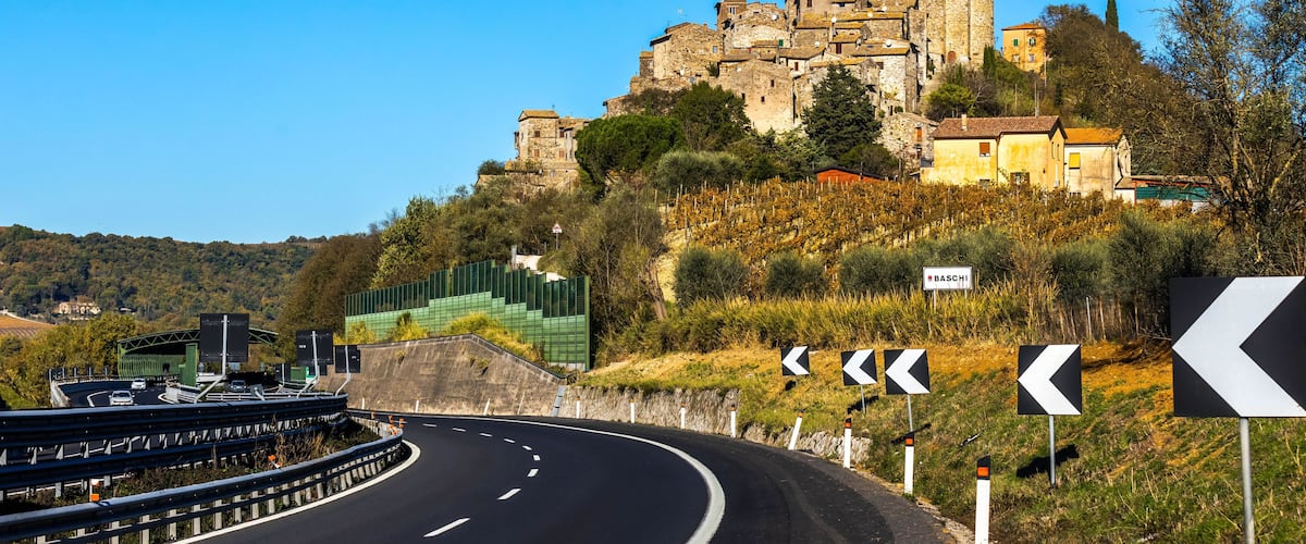 A winding road with directional signs, a historic stone building on a hill, and a clear blue sky.