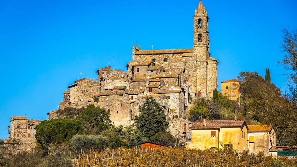 A historic stone building with a bell tower, surrounded by vineyards and trees.